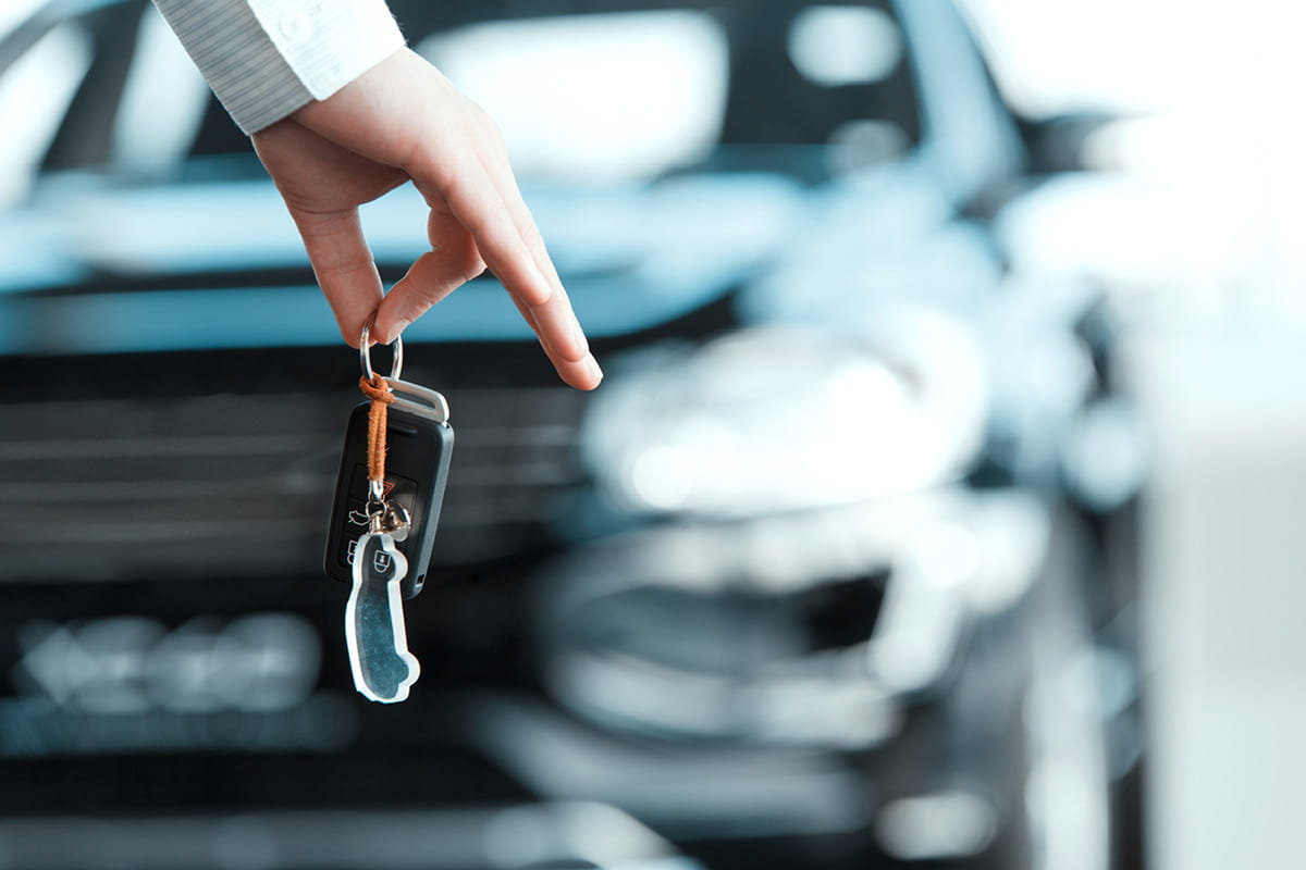 zu sehen ist eine Hand, die einen Autoschlüssel hält, im Hintergrund ist ein Auto